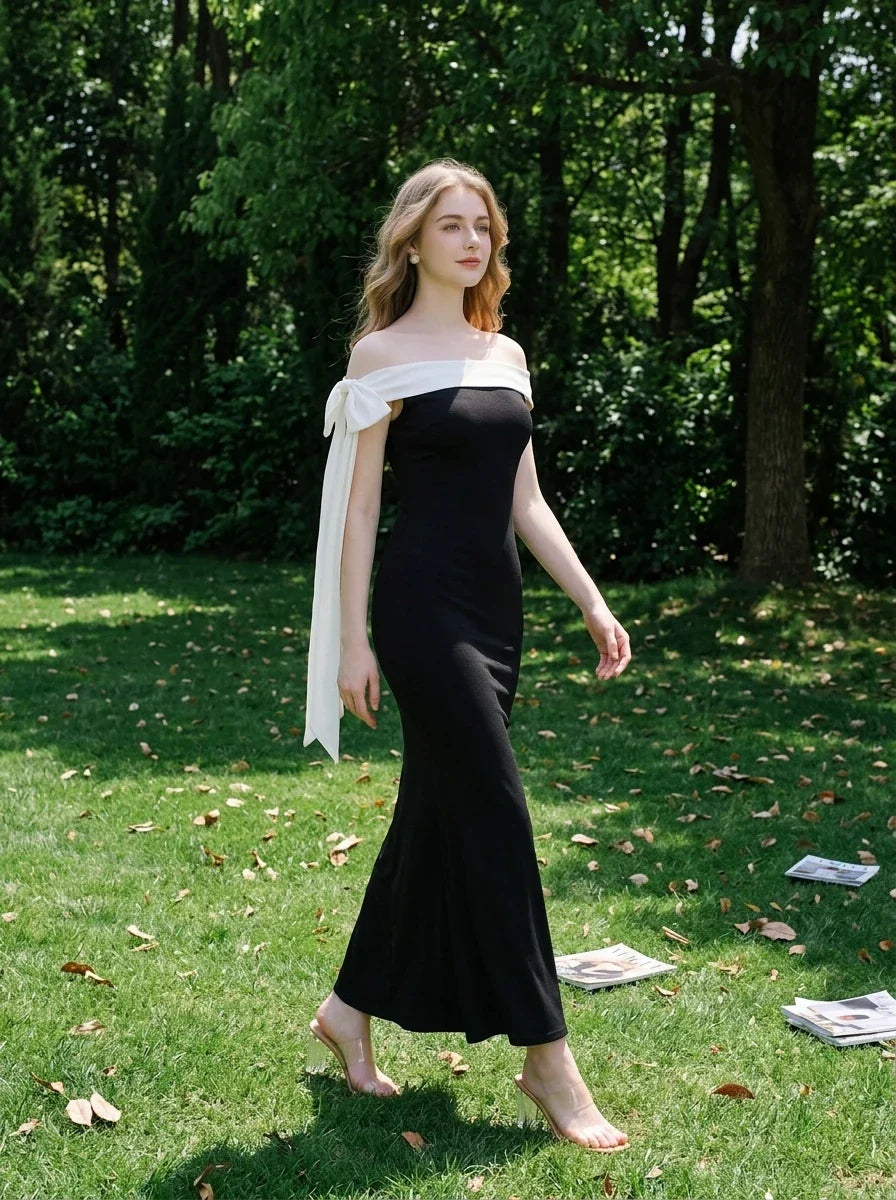 Woman walking in a black off-the-shoulder mermaid gown with a white sash and bow detail in a grassy outdoor setting.