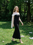 Woman walking in a black off-the-shoulder mermaid gown with a white sash and bow detail in a grassy outdoor setting.