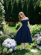 Woman wearing a navy blue off-the-shoulder midi dress with a pleated skirt, posing in a garden with hydrangeas.