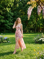 Woman wearing a pink floral print maxi dress with spaghetti straps and a high slit, walking in a sunlit garden with trees in the background.