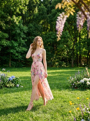 Woman wearing a pink floral print maxi dress with spaghetti straps and a high slit, walking in a sunlit garden with trees in the background.