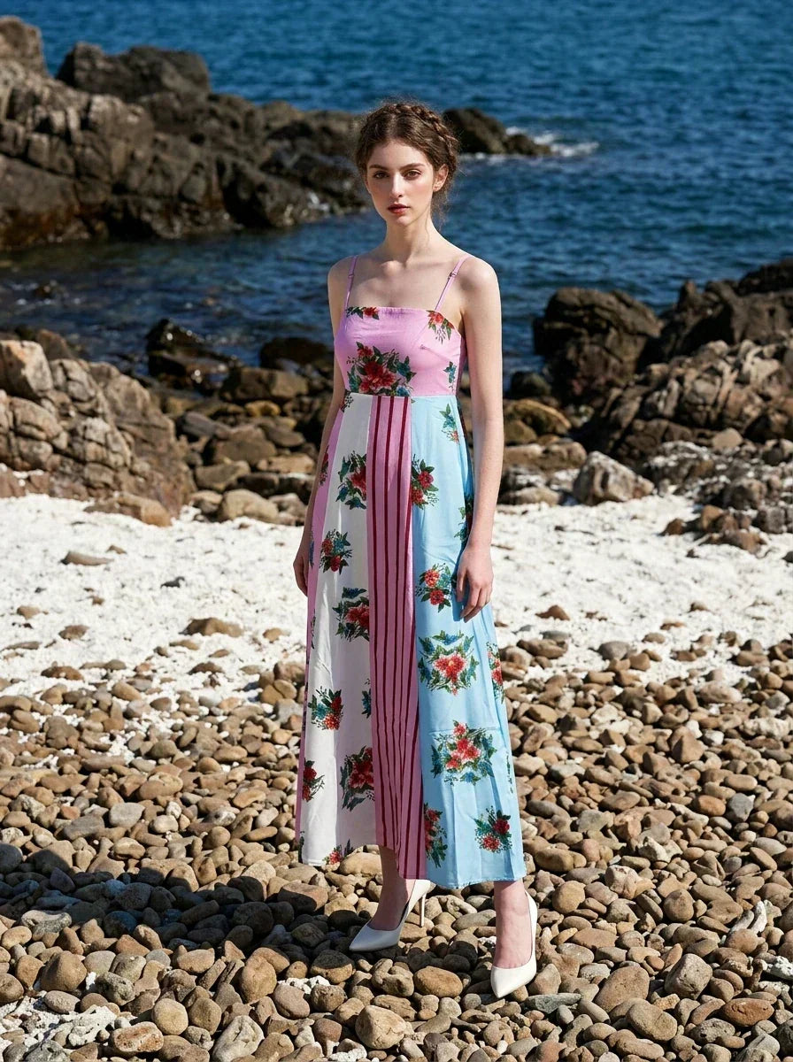 woman in a pink, blue, and white floral print maxi dress with spaghetti straps, posing on a rocky beach with the ocean in the background