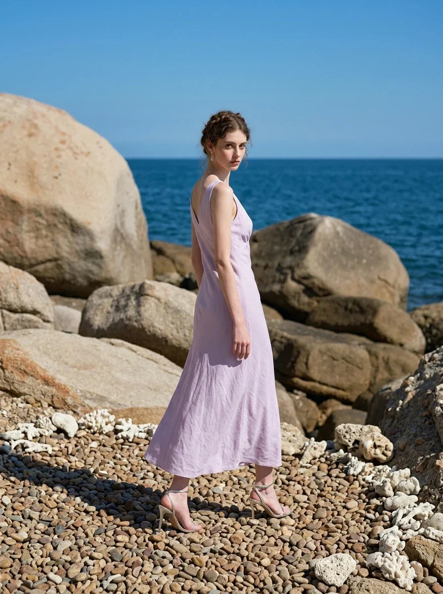 Model wearing a lavender midi dress with a V-neckline and sleeveless design, walking on a rocky beach.
