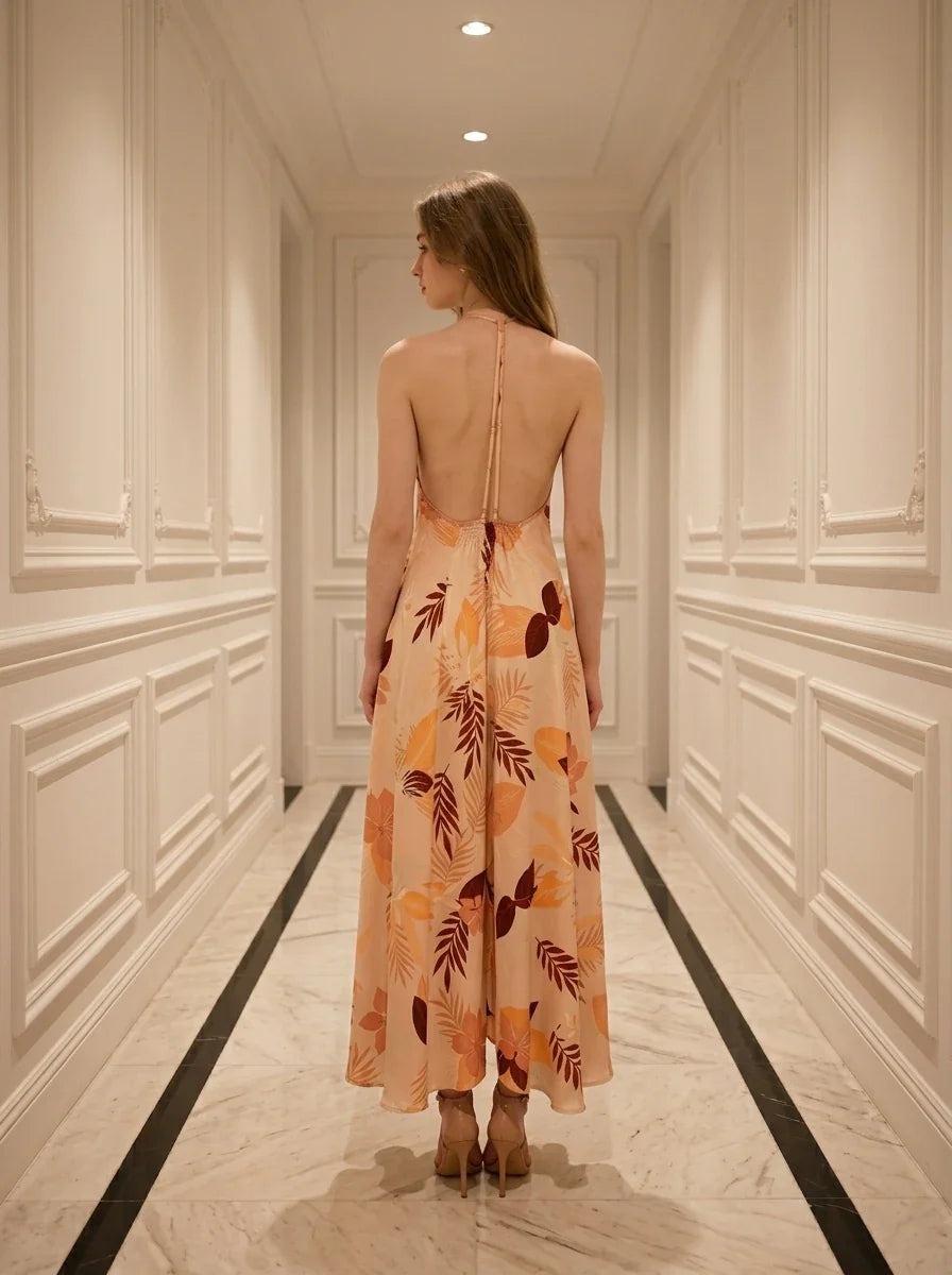 Woman standing in a hallway wearing a long, floral print, halter neck dress with an open back, viewed from the back.
