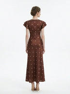Back view of a woman wearing a brown polka dot midi dress with ruffled sleeves and a v-neckline, standing against a white background.