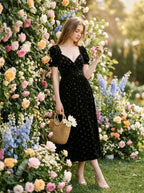 A woman stands next to a floral wall wearing a black velvet midi dress with a floral print and carrying a straw basket.