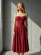 Woman wearing a deep red fit and flare midi dress with spaghetti straps, standing near a window with blinds casting shadows