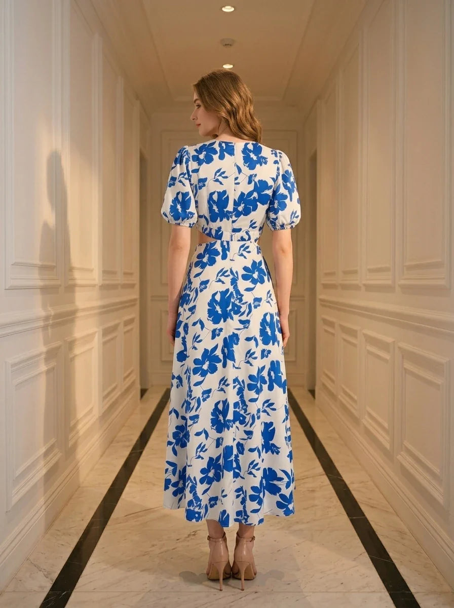 Back view of a woman wearing a white and blue floral print midi dress with cut-out waist and puff sleeves, standing in a hallway.