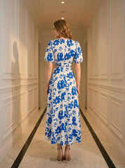Back view of a woman wearing a white and blue floral print midi dress with cut-out waist and puff sleeves, standing in a hallway.