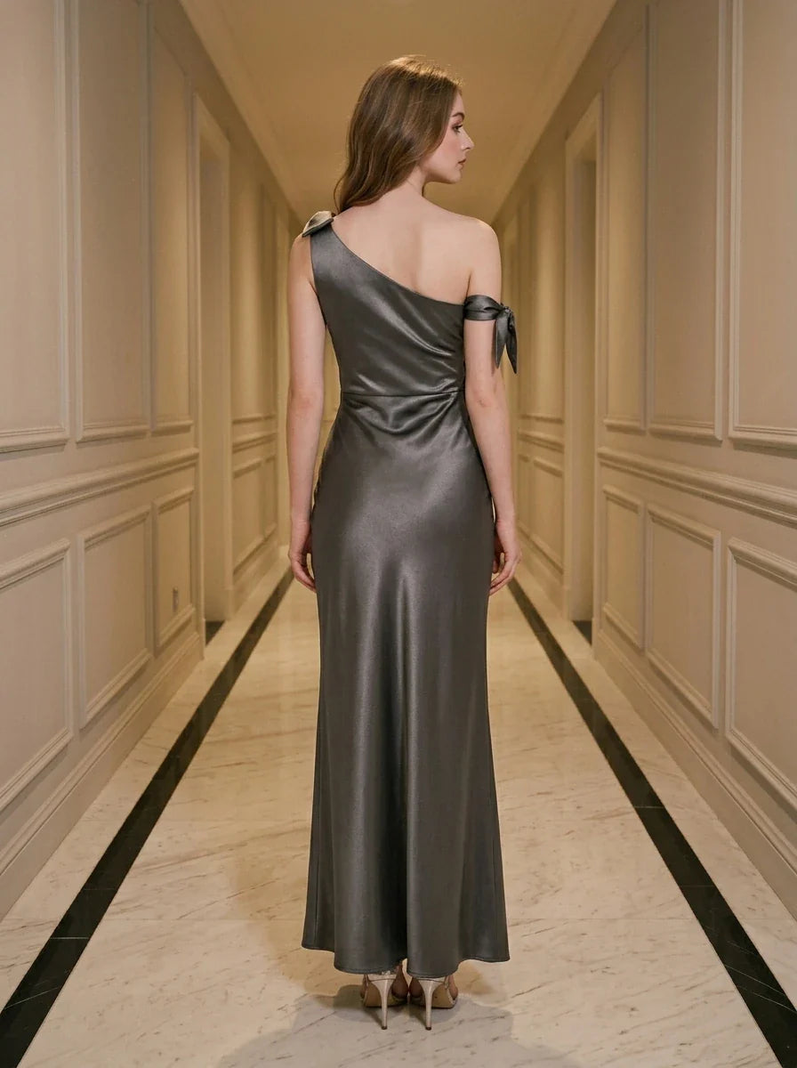 Back view of a woman wearing a floor-length grey satin one-shoulder dress with a high slit and bow accents.