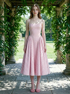 A woman models a pink and white gingham midi dress with a square neckline and a full, pleated skirt, standing in an outdoor garden archway.