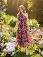 Woman wearing a navy floral print one-shoulder dress with a tiered skirt and ruffle detail, standing in a garden with hydrangeas and flowers.