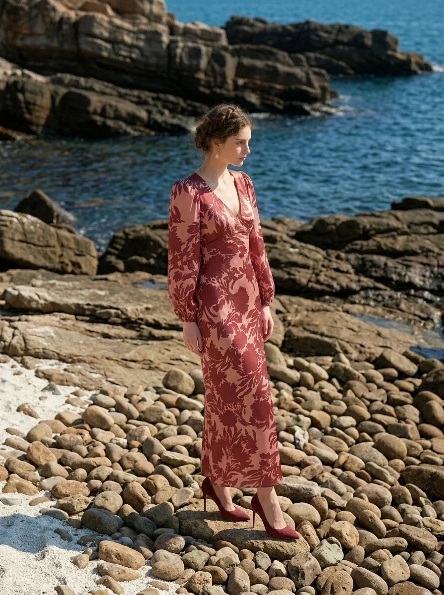 woman wearing a pink and red floral print satin maxi wrap dress with long bishop sleeves and a v-neck, posing on a rocky beach with the ocean in the background