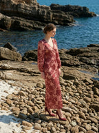 woman wearing a pink and red floral print satin maxi wrap dress with long bishop sleeves and a v-neck, posing on a rocky beach with the ocean in the background