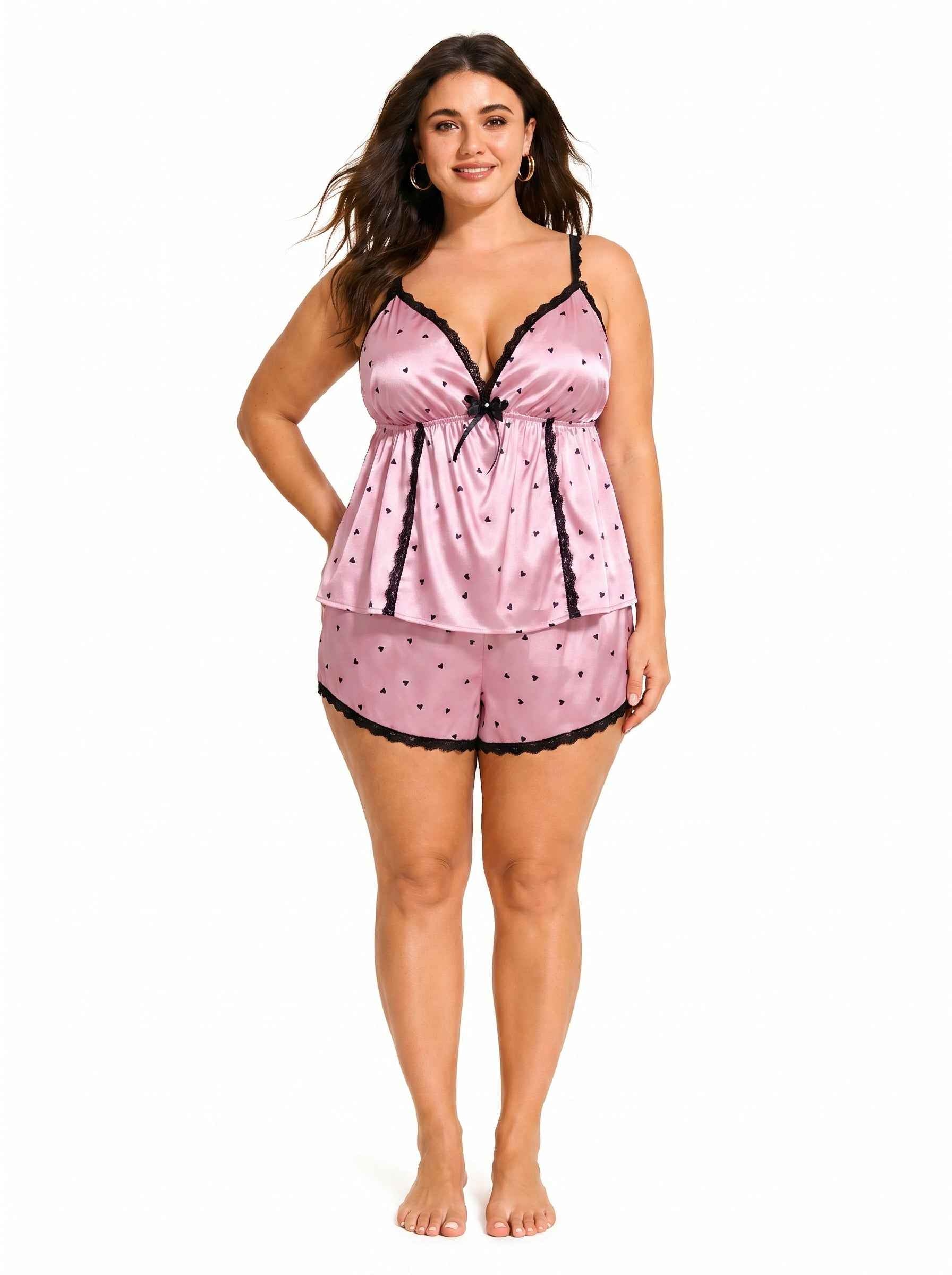 Model wearing a pink satin cami and shorts pajama set with black heart print and lace trim.
