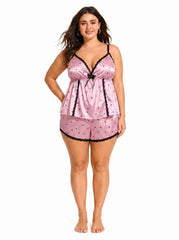 Model wearing a pink satin cami and shorts pajama set with black heart print and lace trim.