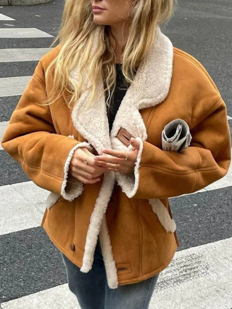 Woman wearing a brown suede jacket with white shearling trim and collar, standing on a crosswalk, holding a newspaper.