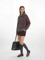 Side profile of a woman in a grey plaid knit jacket, brown mini skirt, and grey knee-high socks, carrying a black handbag.