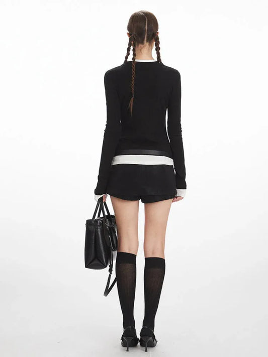 Back view of a woman wearing a black long-sleeved top with white trim, black shorts, black knee-high socks, and black heels, holding a black handbag.