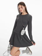 Woman in a dark grey long-sleeve mini dress with a gathered waist, white-trimmed grey socks, and silver shoes, winking and holding a silver handbag and a small striped pouch around her neck.