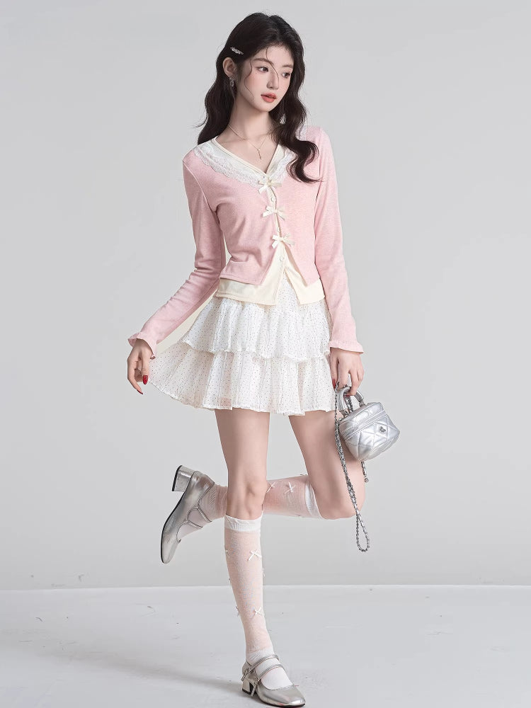 Full body shot of a woman wearing a pink V-neck knit cardigan with lace trim and bow closures, a white polka dot ruffled skirt, knee-high socks, and silver shoes.