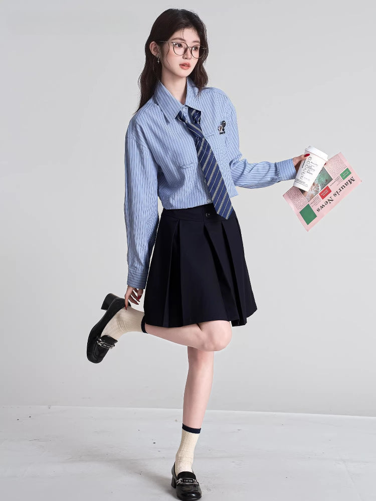 Full body shot of a model wearing a light blue striped button-down shirt with a navy striped tie and a black pleated skirt, holding a coffee cup and a newspaper, with one leg raised.