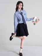Full body shot of a model wearing a light blue striped button-down shirt with a navy striped tie and a black pleated skirt, holding a coffee cup and a newspaper, with one leg raised.