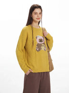 Front view of a woman in a yellow knit sweater featuring a teddy bear applique and brown pants.