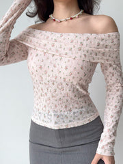 Woman wearing a pink floral off-the-shoulder long-sleeved top with a pearl and rose necklace, paired with gray trousers.