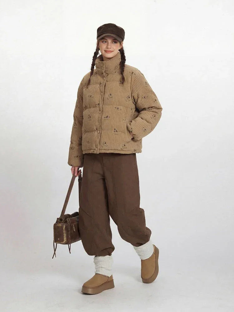 Woman wearing a tan corduroy puffer jacket with a repeating dog print, brown wide-leg pants, and a brown cap, smiling.