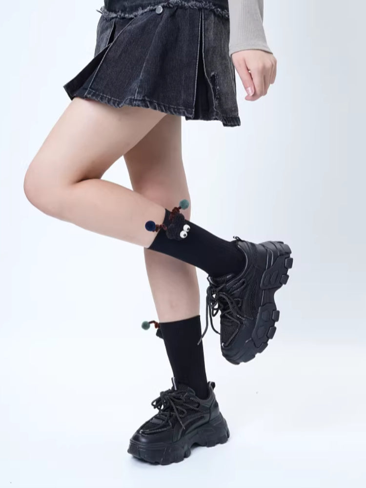A person wearing a dark denim pleated skirt, black crew socks with a black creature applique, and black sneakers.