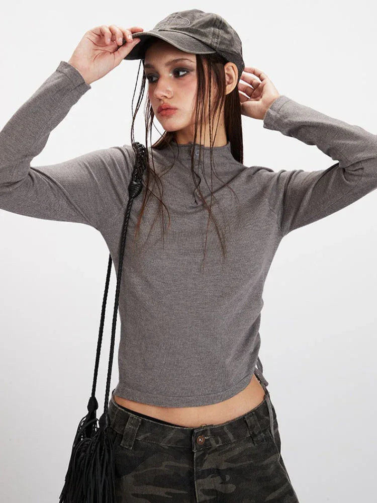 Close-up of a woman wearing a grey long-sleeved mock neck top with button detailing and a cropped hem, paired with camouflage print wide-leg cargo pants, and a grey baseball cap, looking to the side.