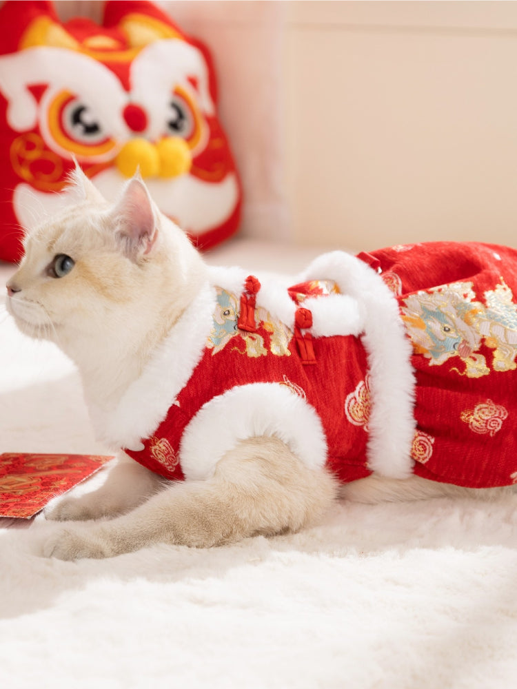 A side view of a cat lying down, wearing a red, festive dress with dragon patterns and white faux fur trim.