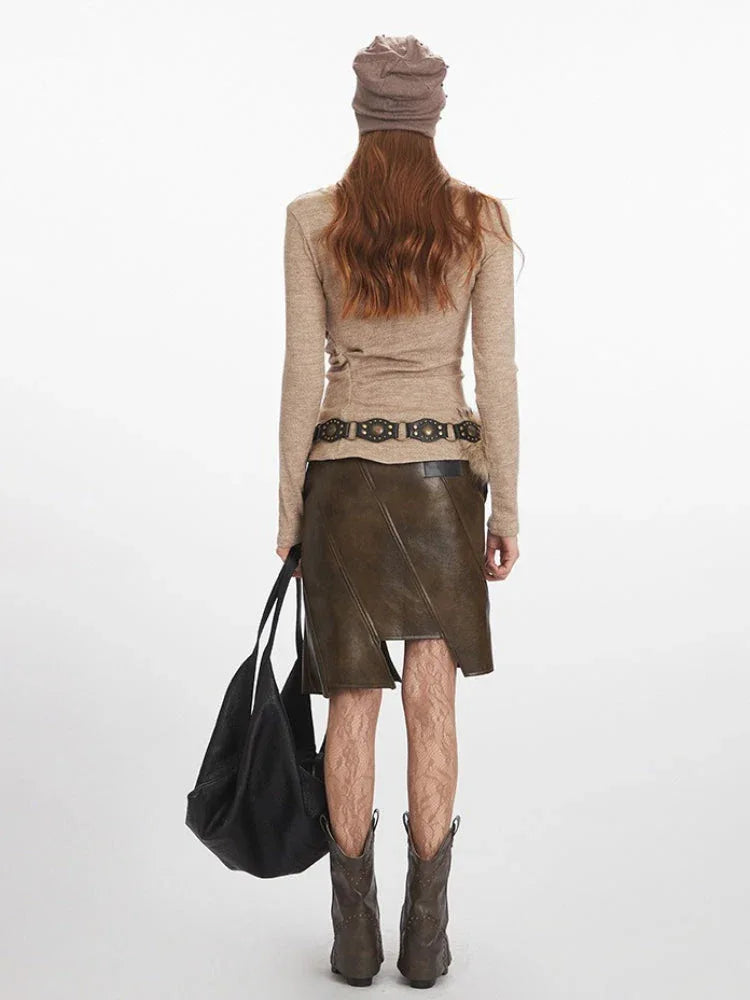 Back view of a woman wearing a beige long-sleeve knit top, an olive green faux leather skirt with an asymmetrical hem, a patterned belt, and brown boots, holding a black tote bag.