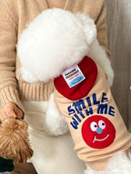 A person holding a white Bichon Frise dog wearing a yellow hoodie with a red hood, while a smaller brown dog looks on.