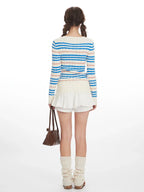 Back view of a woman wearing a striped v-neck sweater with a white ruffled skirt and beige leg warmers, holding a brown handbag.