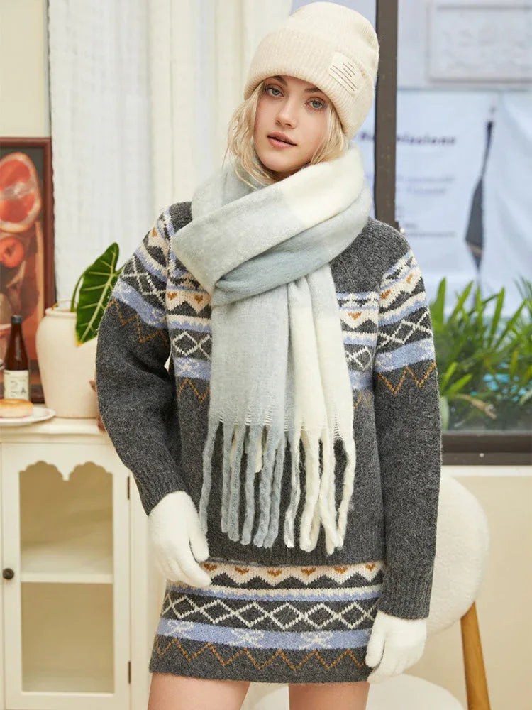 Full body shot of a woman wearing a gray knit sweater dress with Fair Isle pattern, cream beanie, white gloves, and multi-colored fringed scarf.