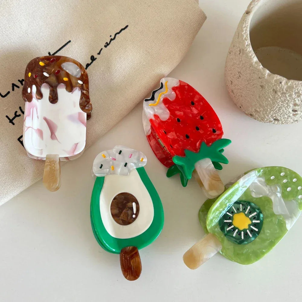 Various fun hair clips shaped like food items, including an ice cream bar and a strawberry, laid out on a textured fabric.