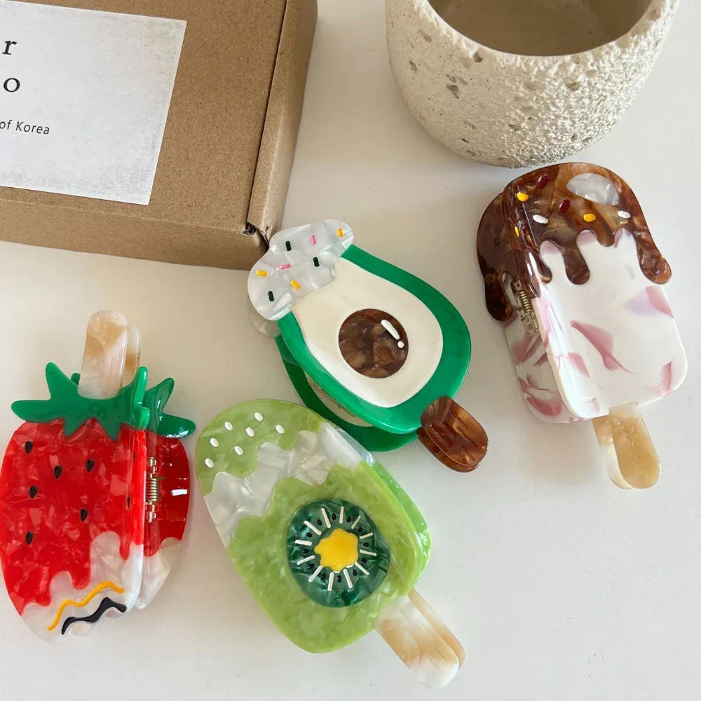 Assortment of fruit and ice cream themed hair clips on a white table, with a box and pot in the background.