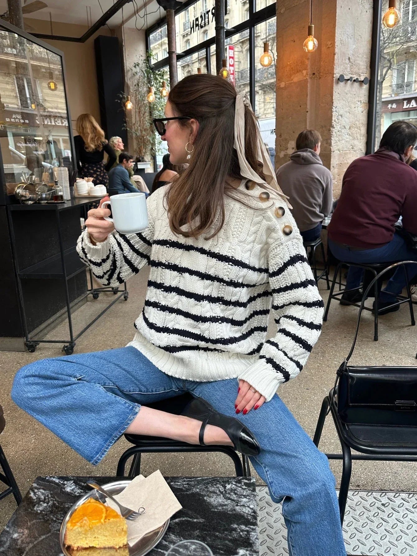 Woman holding a coffee cup while sitting at a cafe table, wearing a striped knit sweater and blue jeans.