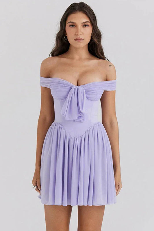 Woman wearing a lavender off-the-shoulder mini dress with a draped bodice and pleated skirt, front view.