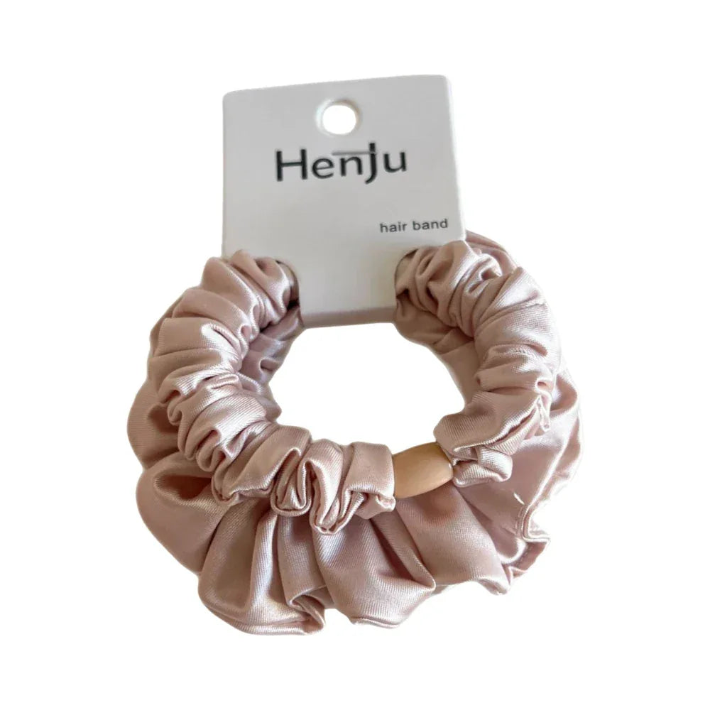 A close-up shot of a light pink satin scrunchie with a wooden bead detail, attached to its product card. The scrunchie has a ruffled texture.