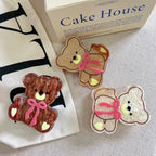 Three teddy bear hair clips in brown, beige, and cream colors, arranged on a textured surface with a box and bag in the background.