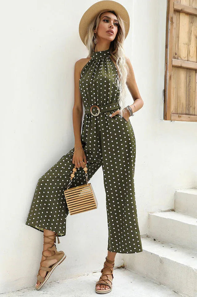 Woman wearing a green polka dot halter neck wide leg jumpsuit with a belt, straw hat, and wooden handbag.