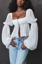 Woman with dark curly hair wearing a white cropped blouse with a square neckline and sheer bell sleeves
