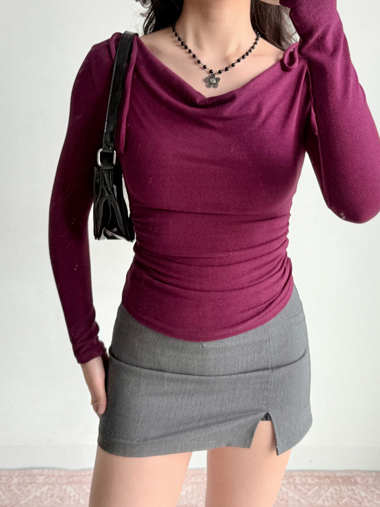 Model wearing a plum long-sleeve top with a draped neckline and ruched sides, paired with a grey skirt and handbag.