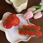 A top-down view of two tomato-shaped hair claws presented on a decorative white plate with a handwritten note and pink tulips.