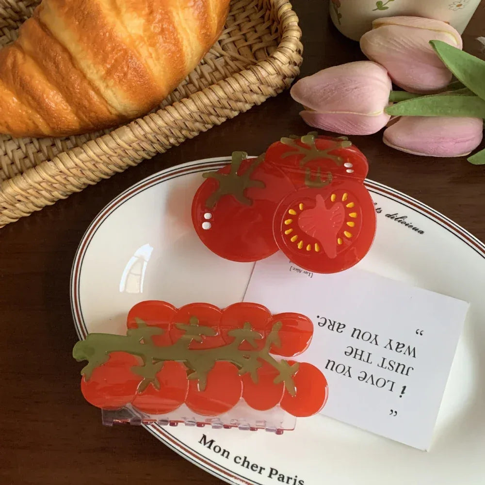 A flat lay image displaying two realistic tomato hair accessories on a white plate, with a croissant and tulips adding to the scene.