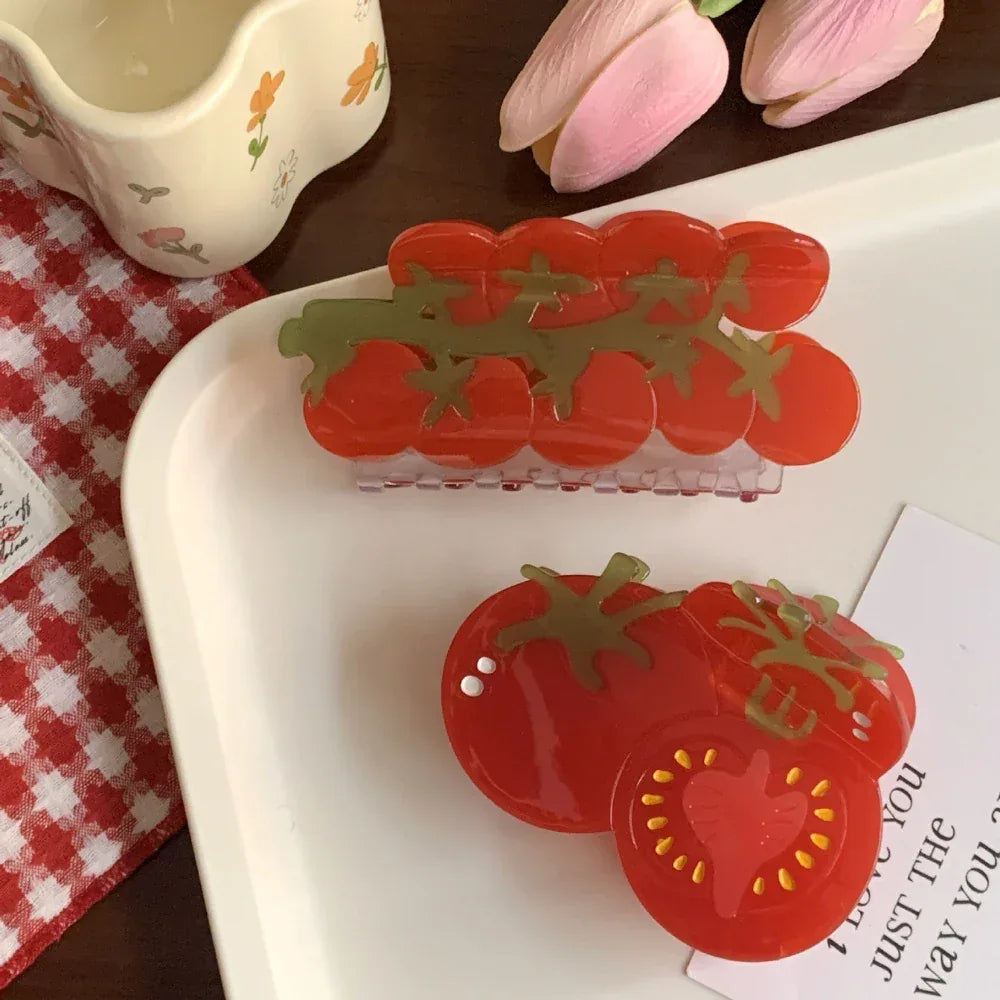 A flat lay composition featuring two tomato-themed hair claws on a white tray, alongside a teacup and pink tulips.