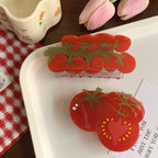 A flat lay composition featuring two tomato-themed hair claws on a white tray, alongside a teacup and pink tulips.